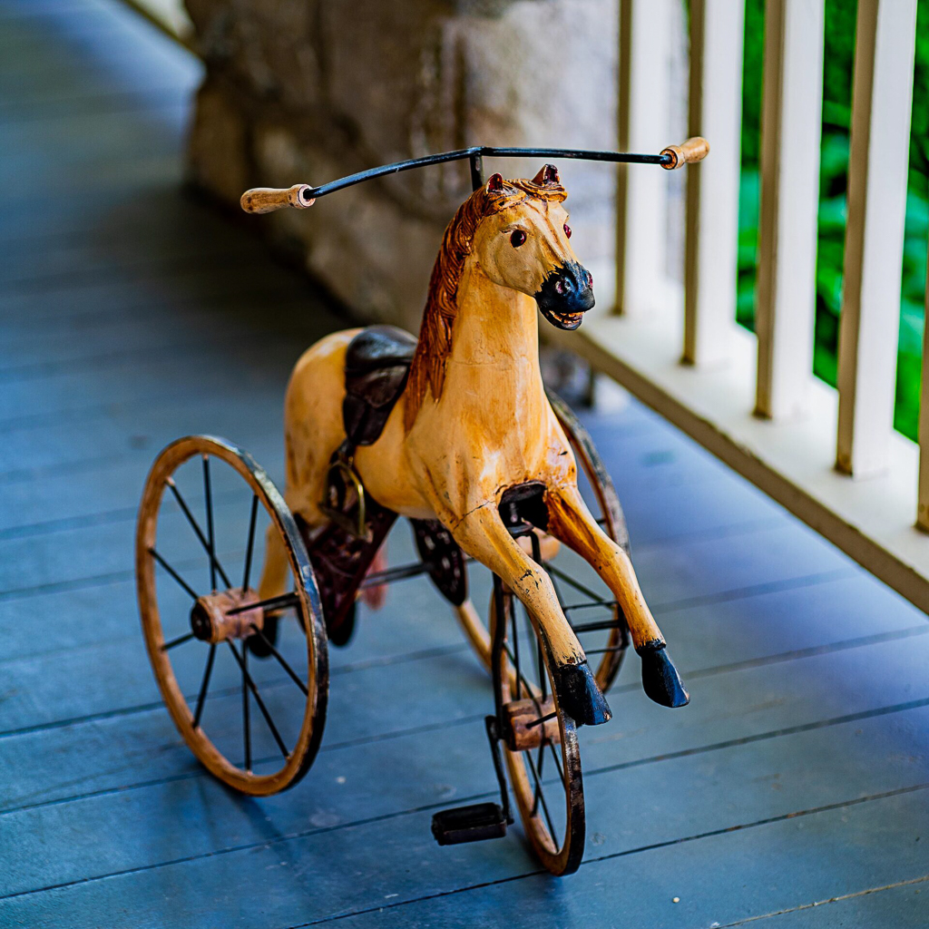 Antique Victorian toy horse on wheels with carved wooden figure and metal spoke wheels, displayed on the painted porch
