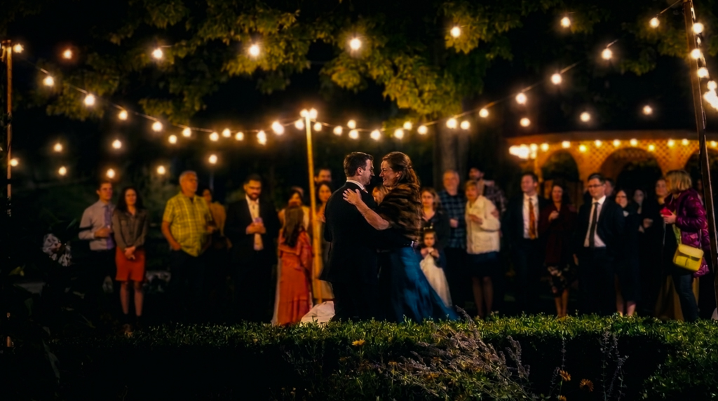 Couple sharing a first dance outdoors under string lights at night, surrounded by guests