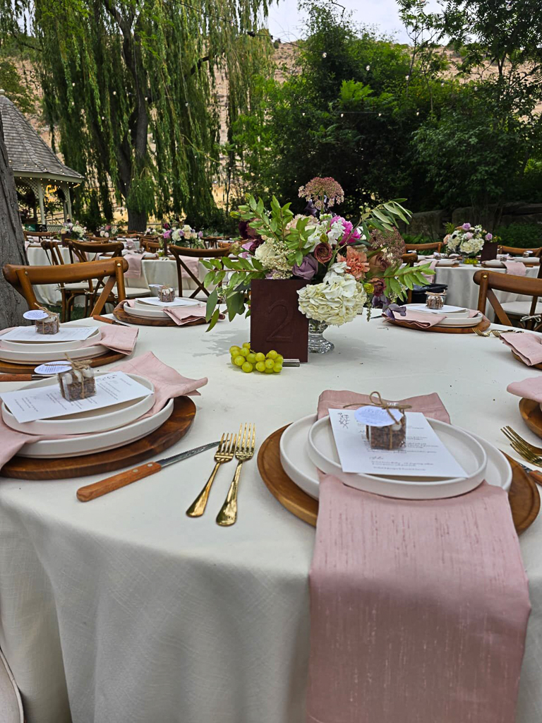 Outdoor wedding reception table set with pink napkins, gold flatware, wooden charger plates, and a lush floral centerpiece